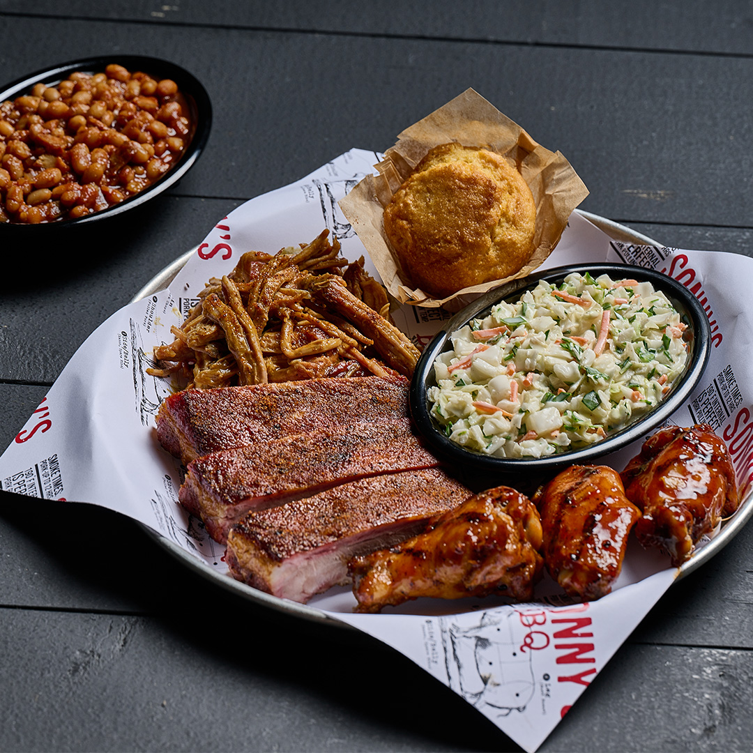 Pitmaster BBQ Tour plate on a table featuring Carolina Pulled Pork, Memphis Dry-Rubbed Ribs, Florida Smokin' Citrus Wings, BBQ Beans, Coleslaw and Cornbread.